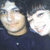 Two people in dark clothing pose closely together, one with a neutral expression and the other smiling broadly across four -like