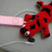 Red cartoon character with black spots and white eyes, holding a pink object against gray background.