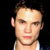 Static pixelated portrait of a person with light skin, dark hair styled upward, and visible facial features including eyes, nose, mouth, ear, and neck against a black background.