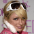 Pixelated image of a person with light brown hair wearing dark sunglasses on their head, a white collar, and a pink garment with a red tie, set against a light gray background with purple accents.