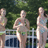A group of women in swimsuits stand behind a white fence with green foliage background, some holding items.