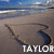 Beach scene with blue sky, ocean waves, and sandy shore featuring a dark curved object on sand and white text reading TAYLOR.