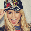 A person with long blonde hair wearing a black and white striped cap with red and yellow floral patterns, holding the cap with their right hand while sticking out their tongue.