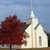 Pixelated image of a white church with a tall steeple, dark roof, and single vertical window, next to a large red tree under a light blue sky.