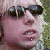 Pixelated close-up of a person wearing dark sunglasses with a light-colored background and green foliage on the right.