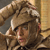Pixelated image of a person with light brown hair wearing a dark cap, holding a small object near their face against a blurred background.