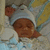 A baby wearing a white knit hat with ear flaps lies on a yellow and blue patterned blanket, eyes closed, with a slightly downturned mouth.