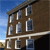 Pixelated image of a brown brick building with multiple windows, a dark roof, and a white horizontal stripe against a blue sky background.