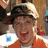 Pixelated image of a person wearing an orange shirt and dark cap, with visible red mouth area and blue square on white background.
