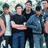 Group of young men in casual clothing standing outdoors with arms crossed, some wearing dark jackets and blue jeans.