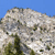 Pixelated static image of a large light-colored rock formation with patches of green vegetation against a solid blue sky background.