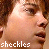 Pixelated close-up of a person's face with light skin tone, brown hair visible on left side, and white text reading 'sheckles' at bottom.