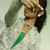 A person with dark hair and light skin holds a clear bottle with green liquid, hand raised to forehead in a saluting gesture against a pale background.