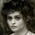 A grayscale pixelated portrait of a person with curly hair, wearing a light-colored top, against a dark background.