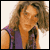 Pixelated static image of a person with long curly hair wearing an orange top and purple vest against a light background.