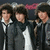 Group of four people in dark coats with white collars posing together against a dark background with red text.