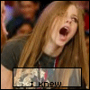 A woman with long light brown hair in a dark gray shirt, mouth open wide as if shouting or singing, against a blurred crowd background.