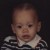 Pixelated image of a child's face with light skin, dark eyes, and short hair, wearing a white-collared shirt with dark stripes against a dark background.