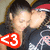 Two people in close physical proximity with one person's face near the other's cheek, wearing dark clothing and a red cap, with a large red symbol containing white numbers on the left side.