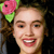 A pixelated close-up of a person with long brown hair wearing a pink flower with green leaves on the left side of their head, smiling with visible teeth and red lips.