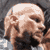 Pixelated static image of a bald human head with brown hair on the sides, dark eyes, and visible facial features against a gray background.