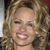 A pixelated close-up of a person with light brown curly hair, wearing a dark top, and smiling with visible teeth.