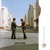 Two figures stand on a paved surface under a light blue sky, one holding a small object near the other's face.