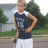 A pixelated static image shows a person standing outdoors with one hand on their hip, wearing a dark sleeveless shirt and light-colored shorts against a blurred background of greenery and pavement.