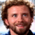 Pixelated close-up of a person with brown curly hair, light skin tone, and visible facial stubble wearing dark clothing against a blurred background.