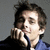 Pixelated static image of a person with dark hair wearing a blue shirt and black jacket, hand near face in contemplative pose against gray background.