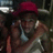 A person wearing a red cap and light-colored tank top, holding a phone to their ear in a dimly lit environment with visible background elements.