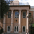 A pixelated image of a classical building with columns, brown facade, dark windows, and a light roof under a blue sky with clouds.