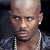 Static pixelated image of a bald man with light skin wearing a silver chain and dark jacket, set against a dark background.