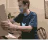 A man in a dark blue shirt with a light blue logo sits at a desk, performing sequential hand and facial gestures against a beige wall background.