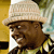 Pixelated image of a person wearing a light-colored wide-brimmed hat and a patterned shirt, with a blurred background featuring yellow and green tones.