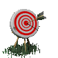 A circular target with red and white concentric rings, an arrow embedded in the center, mounted on three wooden legs above green grass.