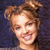 Pixelated close-up of a smiling person with light brown hair styled in a high bun against a dark blue background.
