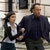 Two people stand side-by-side in a pixelated image; the person on the left wears a dark jacket over a light shirt and dark pants, while the person on the right wears a dark long-sleeve shirt and dark pants.