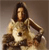 Pixelated image of a person with long dark hair wearing a light-colored top with a dark circular mark on the chest, seated with arms resting on a surface.