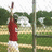 A pixelated figure in a red shirt and white shorts stands on a grassy field with trees and a pole in the background.