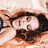 A person with dark hair lies under a light-colored blanket, resting on an orange patterned surface while holding their head with one hand.
