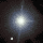 Pixelated starburst with central bright core and radiating light rings on dark background, featuring small white square element.