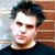 Pixelated portrait of a person with dark hair against a brick wall background, wearing a dark top.