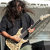 Pixelated image of a person with long dark hair wearing a dark shirt, holding a light-colored electric guitar against a blurred background.