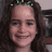 A person with long dark hair and a green headband smiles, wearing a light-colored top against a blurred background.