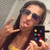 A pixelated figure with long dark hair wears sunglasses and holds a small device with colored buttons while making a hand gesture.