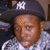 A pixelated close-up of a person wearing a dark cap with white 'NY' lettering and a light checkered shirt, set against a blurred background.