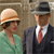 Two figures in vintage attire stand side-by-side against a textured brown background; one wears a light-colored hat and green dress, the other a dark suit with a red tie and gray fedora.