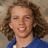 Pixelated portrait of a person with curly brown hair wearing a blue top, smiling with mouth open showing teeth against a neutral background.