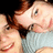 Two people in close proximity, one with brown hair and light blue clothing, the other with dark hair, both smiling with visible teeth.
